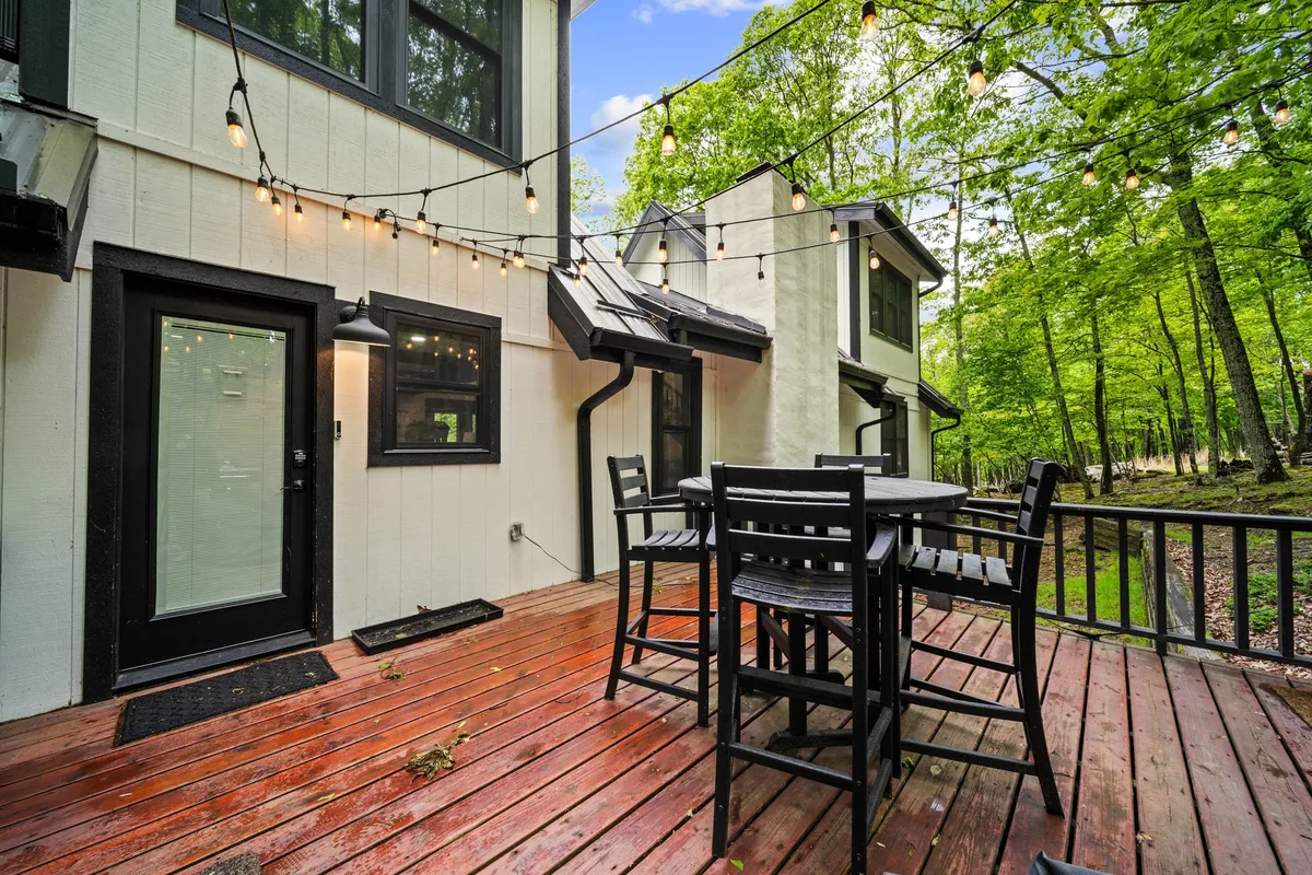 An inviting wooden deck set amidst lush trees, featuring a spacious outdoor dining table and string lights overhead. The setting suggests a seamless blend of indoor comfort and outdoor enjoyment, perfect for gatherings, meals, and soaking up the mountain atmosphere.