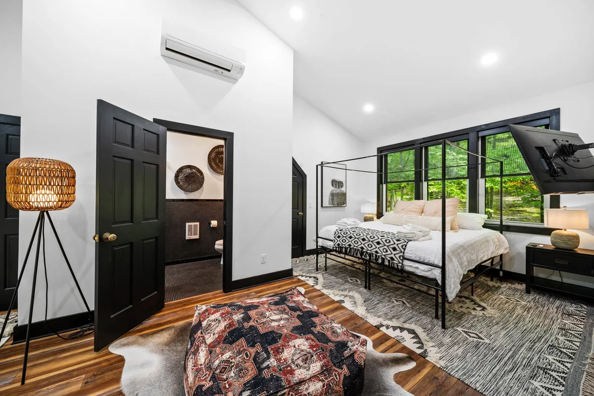 A serene bedroom with a canopy bed, large windows that let in abundant natural light, a decorative rug, and bedside lamps. The room features calming colors and soft linens, with views of the surrounding landscape visible through the windows.