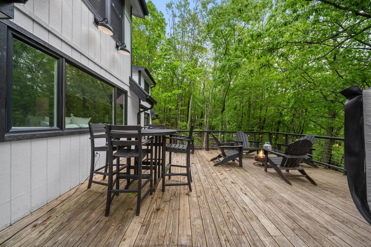 A broad deck set with a large dining table and high chairs, as well as Adirondack chairs, is surrounded by greenery, offering space for outdoor meals or casual relaxation in nature.