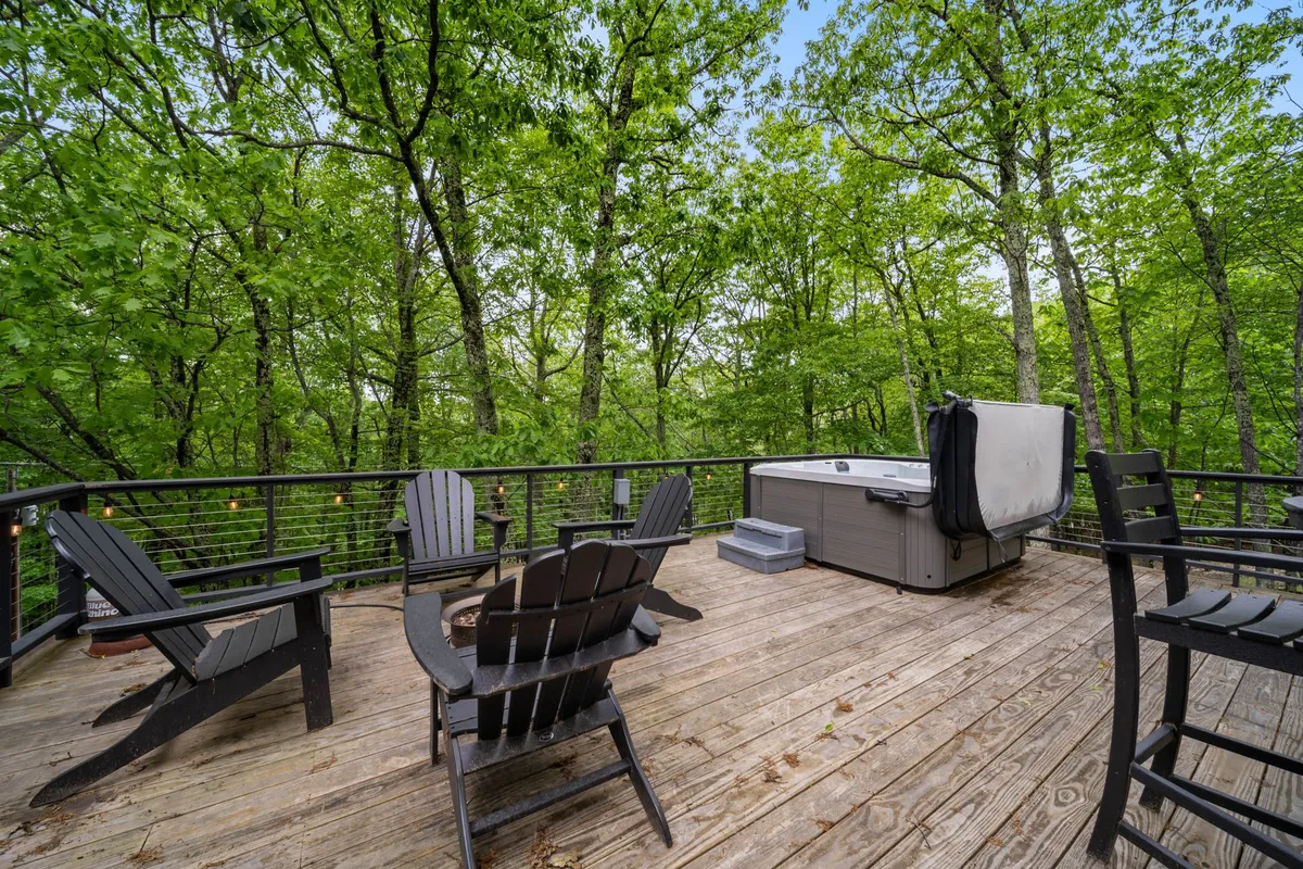 A bubbling hot tub sits on a wooden deck amidst the trees, accompanied by Adirondack chairs and natural surroundings. The peaceful setting invites guests to soak while enjoying the crisp mountain air and tranquil scenery.