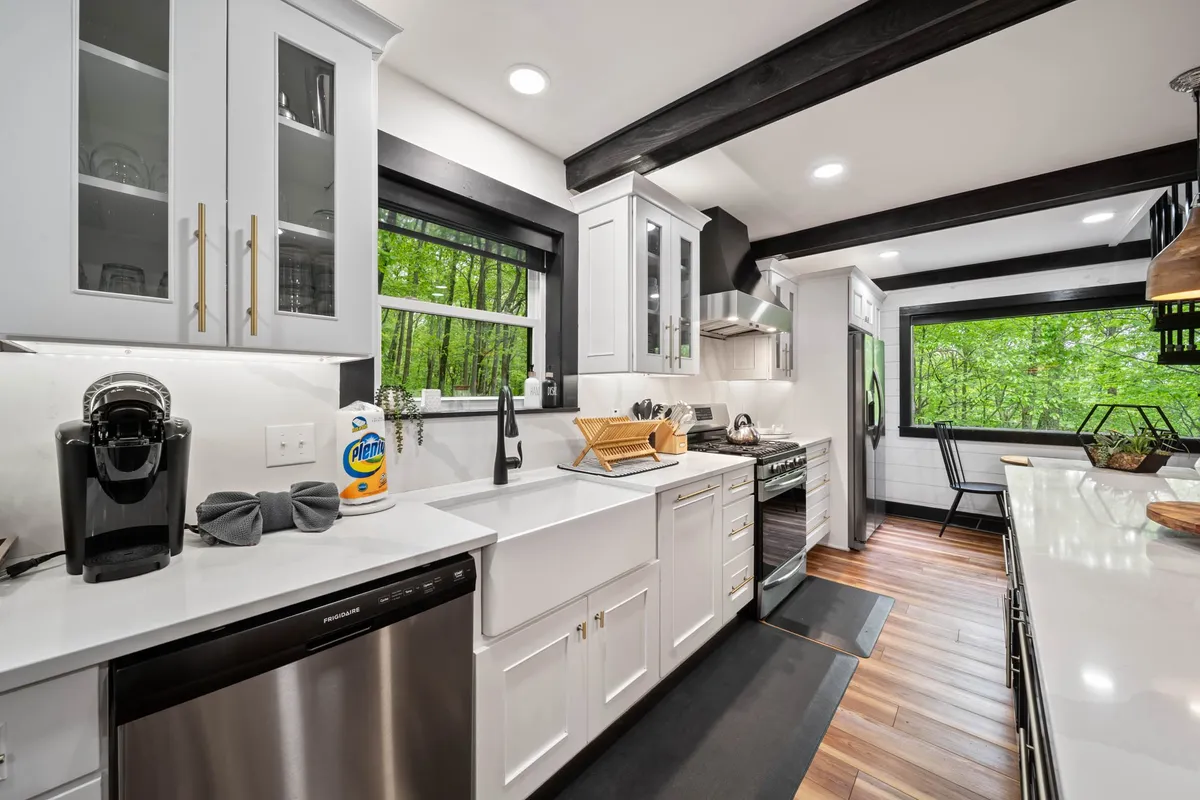 A bright, modern kitchen filled with natural light, featuring sleek stainless steel appliances, large windows with outdoor views, and white custom cabinetry. The kitchen has a functional layout perfect for group cooking and meal prep.