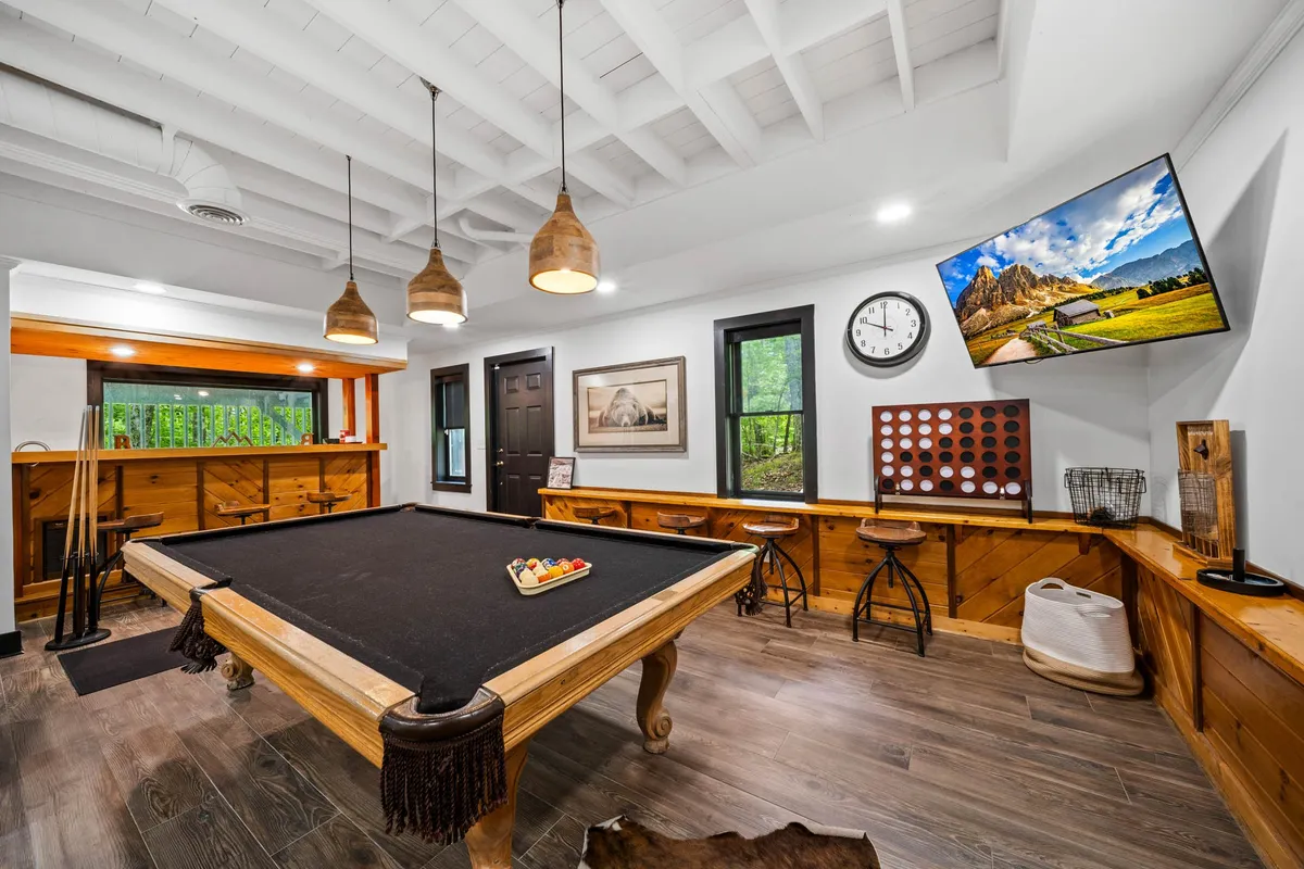 The billiards/game area features a sleek pool table beneath modern pendant lighting, complemented by a wall-mounted TV and a bar area in the background. This setup invites casual competition and socializing, making it ideal for game nights or celebrations with friends.