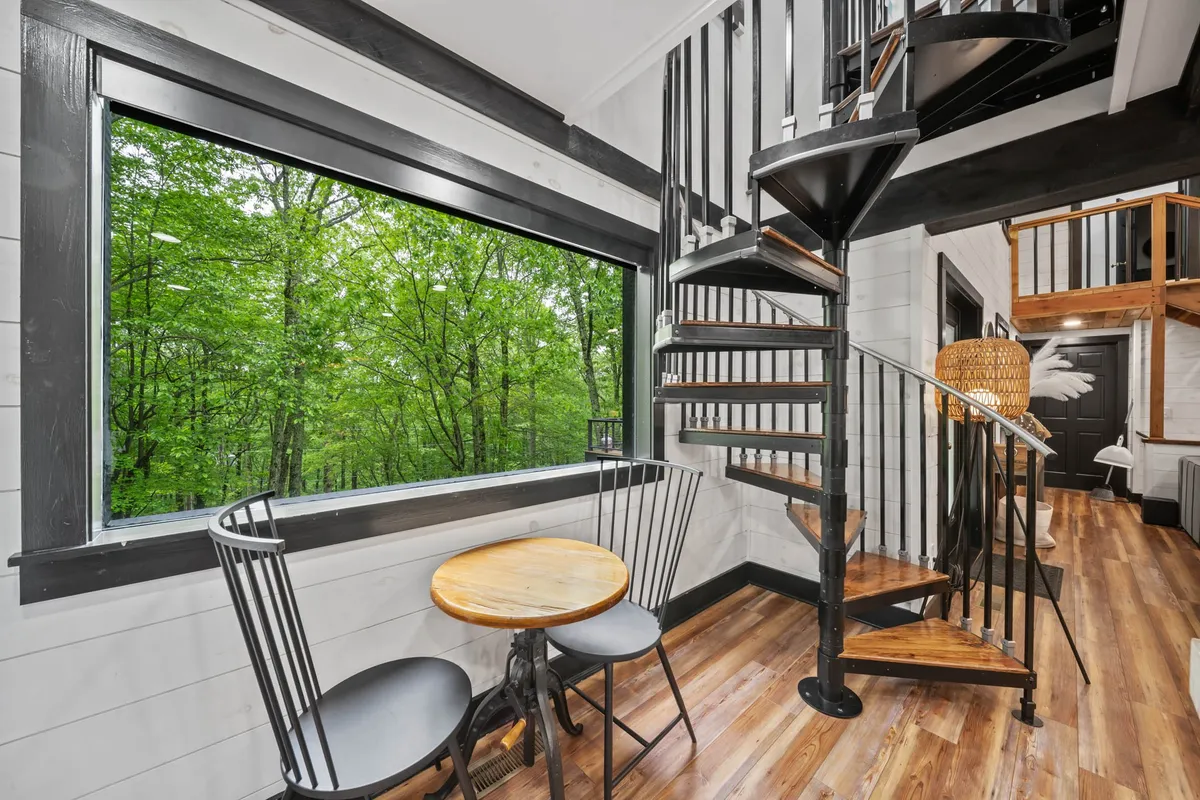 A tranquil interior scene featuring a large window that overlooks lush greenery outside. The spiral staircase and round wooden table create a cozy corner, and the abundant natural light suggests a strong visual connection to the outdoors. The setting feels perfect for enjoying a morning coffee while taking in the surroundings.