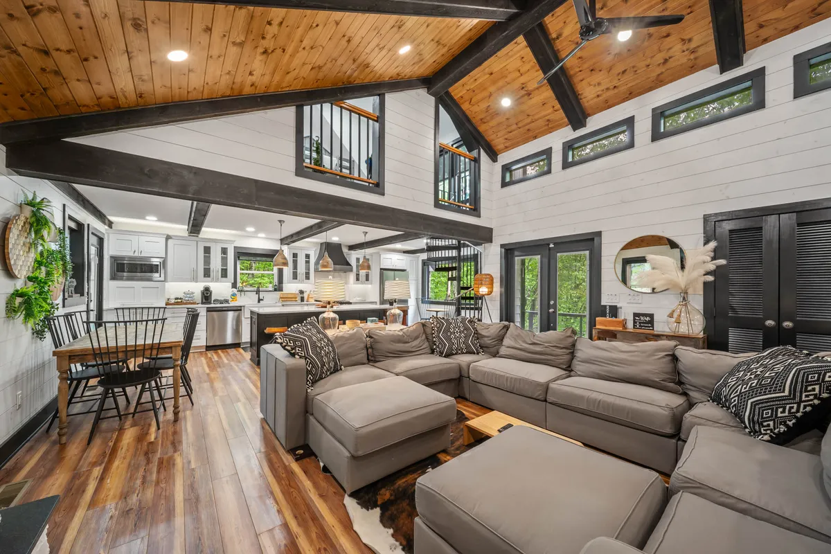 The image showcases a spacious, inviting interior with a large sectional sofa centered in an open-concept living space, exposed wooden beams overhead, a modern kitchen in the background, a cozy dining area, and touches of greenery from decorative plants. The scene exudes warmth and comfort, ideal for gatherings and relaxation.