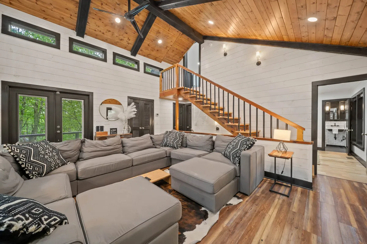 An expansive open-concept living space with a large sectional sofa, staircase, and dramatic floor-to-ceiling windows that flood the room with light. Decorative accents and the arrangement of seating make this a natural hub for gatherings, conversation, and enjoying the mountain views beyond.