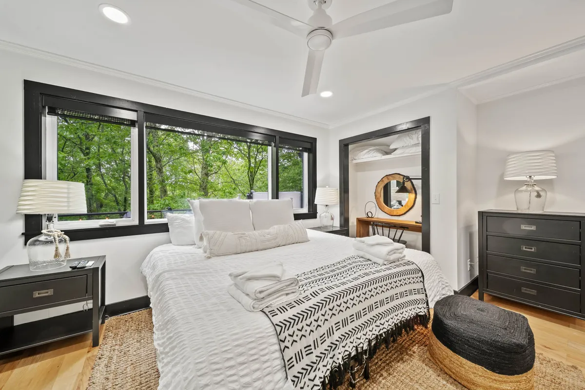 A stylish bedroom featuring a bed adorned with decorative throws, modern bedside lamps, a unique circular mirror, and expansive windows offering unobstructed views of greenery. The space appears tranquil and cozy, perfect for relaxation.
